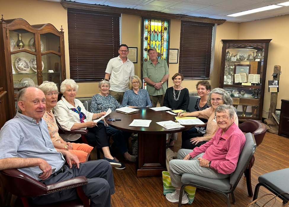 Group of seniors seated around a round table in a room with display cabinets and a stained-glass window behind them.