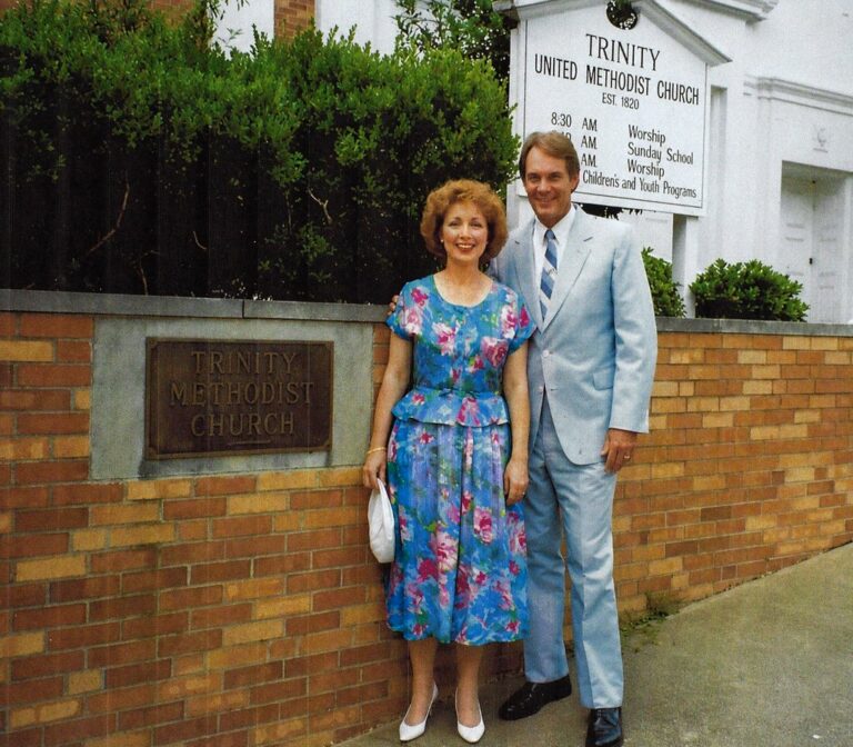 Smiling couple poses outside a brick church; woman in a blue floral dress holds a white purse, man in a light blue suit stands beside her, with a Trinity United Methodist Church sign in the background.