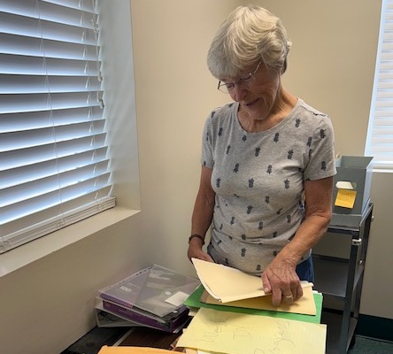 Older woman with short gray hair standing at a desk, sorting through folders and papers by a window.